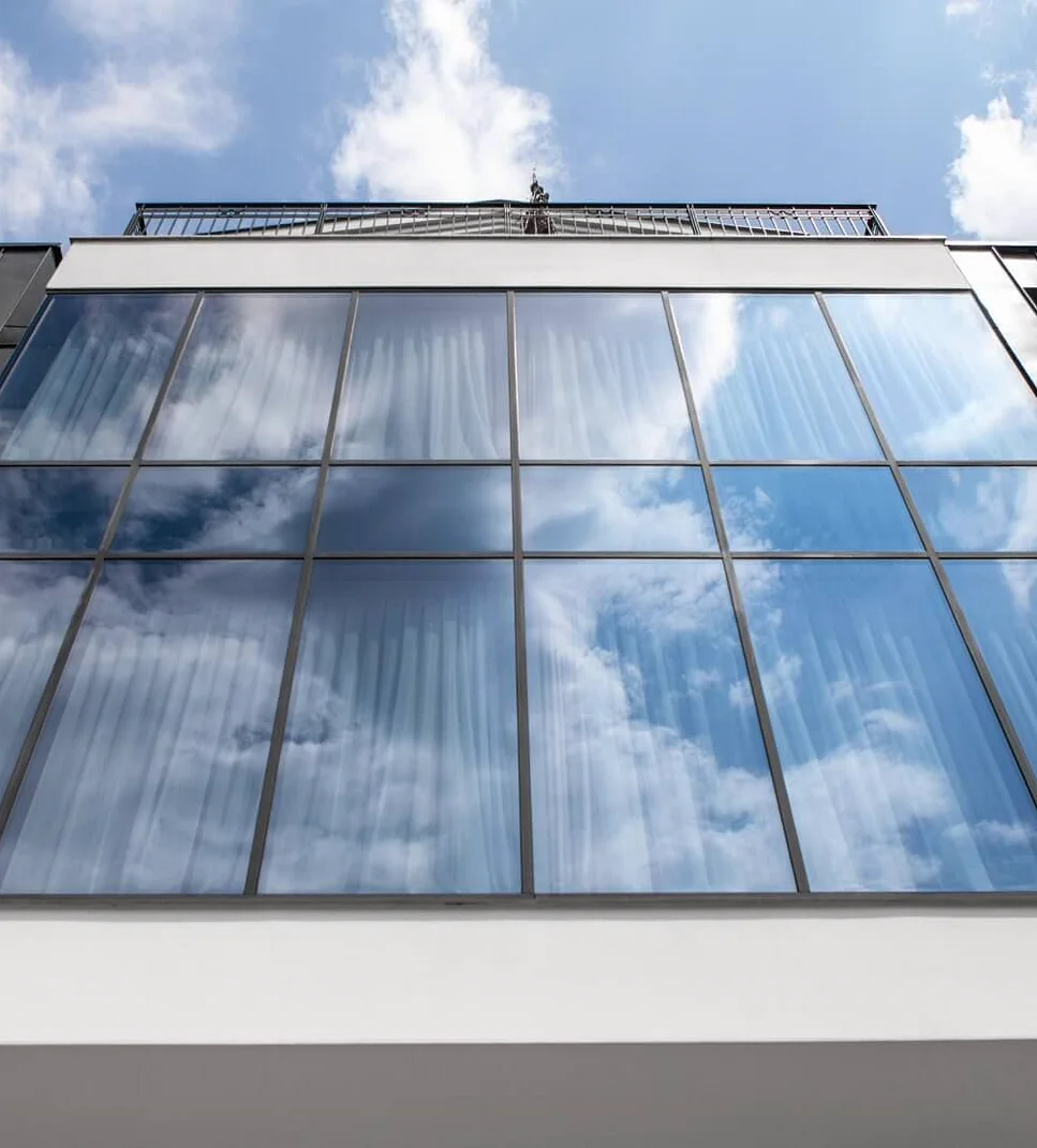 Przeszklona fasada aluminiowa słupowo-ryglowa widziana z dołu — siatka wielkich tafli szkła odbijających niebo z chmurami, biała elewacja i attyka z balustradą